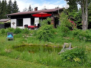 Das Ferienhaus Goldener Hirsch mit Terrasse, rotem Sonnenschirm und kleinem Gartenteich inmitten einer natürlichen Gartenlandschaft